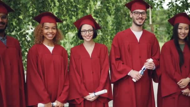 Dolly shot of smiling graduates standing in line on campus, looking at camera and holding diplomas. Gaun yang terang dan papan mortir, pohon dan gulungan terlihat . — Stok Video