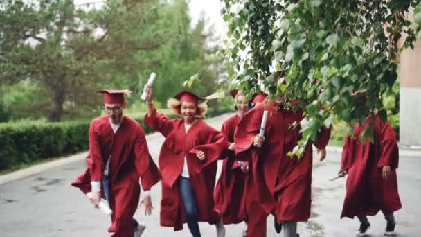 Movimento lento de graduados felizes correndo no campus acenando diplomas e sorrindo vestindo vestidos vermelhos e chapéus. Belas árvores e arbustos são visíveis, está chovendo . — Vídeo de Stock