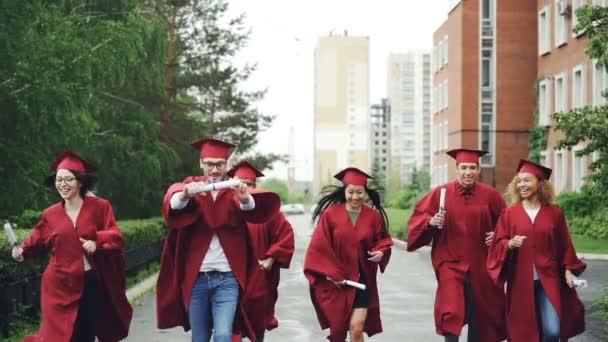 Emocionados estudiantes graduados corriendo a lo largo de la carretera en el campus con diplomas con vestidos de graduación y sombreros. Pequeña lluvia es visible — Vídeos de Stock