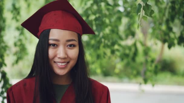 Slow motion portret van aantrekkelijke Aziatisch meisje afstuderen student in toga en mortel halfpension staande op de campus, glimlachend en camera kijken. Jeugd en onderwijs concept. — Stockvideo