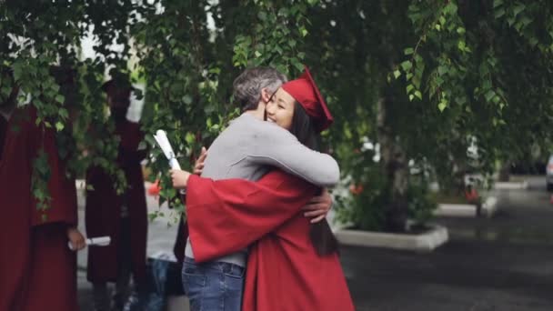 Padre cariñoso está felicitando a su hija en el día de la graduación, la gente está abrazando y riendo al aire libre, mientras que otros graduados son visibles en el fondo . — Vídeo de stock