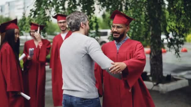 Dumni, że nauczyciel jest gratulacji student, drżenie dłoni i przytulanie go na studiach stojącą na terenie kampusu w tradycyjnych grad odzieży. Koncepcja kształcenia i osób. — Wideo stockowe