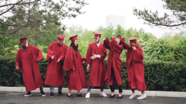 Slow motion of multi-ethnic group of students in gowns and mortar-boards dancing and laughing outdoors on campus on graduation day celebrating event. — Stock Video