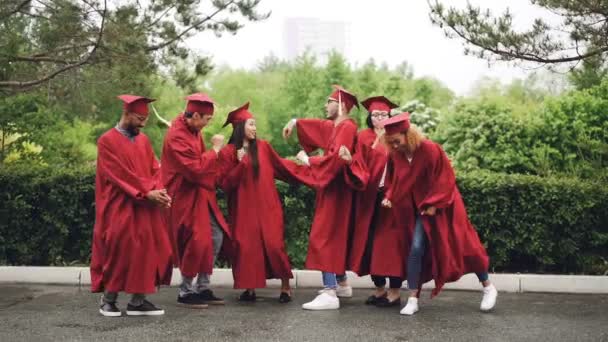 Zwolnionym tempie figlarny dziewczyny i chłopaki studia studentom, taniec, śmiech i zabawa na dzień ukończenia. Piękny letni dzień, zielone drzewa są widoczne. — Wideo stockowe