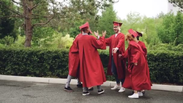 Mouvement lent des jeunes hommes et des jeunes femmes diplômés serrant la main, faisant du high-five, dansant et se câlinant à l'extérieur le jour de gradation avec une belle nature en arrière-plan . — Video