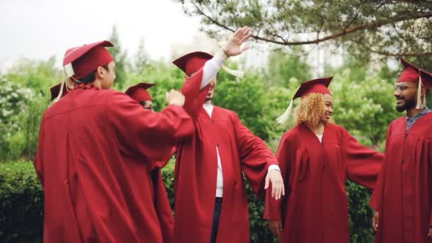 Hombres y mujeres jóvenes emocionados están celebrando el día de la graduación riendo, haciendo chocar los cinco, abrazando y bailando afuera en el campus. La gente usa prendas tradicionales. . — Vídeos de Stock