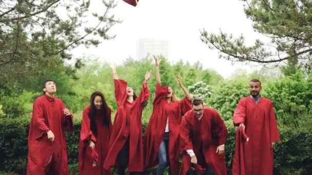 Movimiento lento de graduados felices lanzando morteros al aire, riendo y celebrando la graduación en el campus universitario. Educación, éxito y concepto juvenil moderno . — Vídeos de Stock