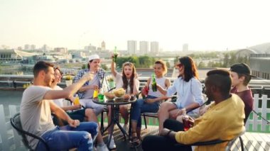 Çekici erkek ve kadın toasting ve gözlük ve şişeler tungur sonra bira ve kokteyl içme ve modern şehir içinde çatıda parti sırasında gülüyor.