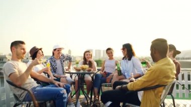 Çok ırklı grup arkadaşıyla konuşurken, kızartma sonra şişe ve gözlük ve içki bira ve meşrubat çatıda masada kımıldıyor. Güzel şehir arka planda görülmektedir.