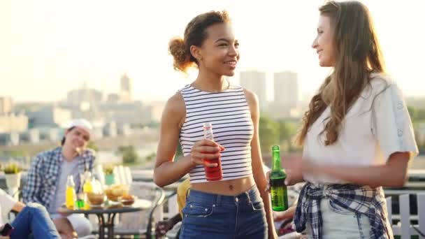 Αρκετά αφρικανικό αμερικανικό κορίτσι μιλάμε για το φίλο του Καυκάσου και πίνοντας κοκτέιλ εκμετάλλευση μπουκάλι κατά τη διάρκεια υπαίθρια πάρτι στον τελευταίο όροφο. Έννοια της φιλίας, της διασκέδασης και της επικοινωνίας. — Αρχείο Βίντεο