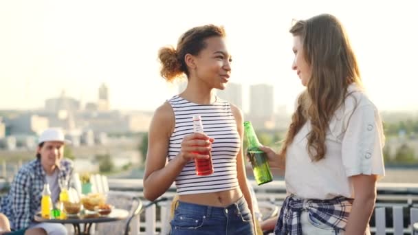 Gelukkig jonge vrouwen zijn chatten, rammelende flessen en drinken staande op het dak met hun vrienden met plezier in de achtergrond. Millennials, vreugde en zomer concept. — Stockvideo