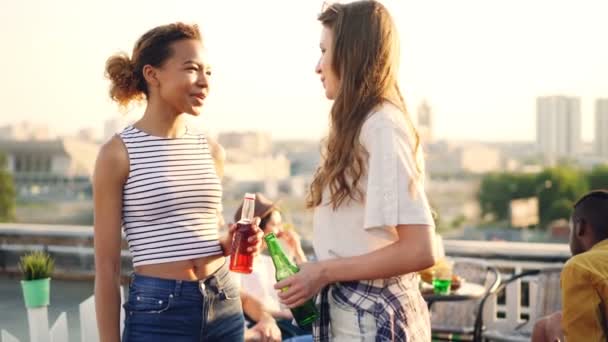 Alegre estudiantes femeninas están hablando, haciendo chocar cinco, tintinear botellas y beber durante la fiesta en la azotea con amigos. Conversación, bebidas y concepto divertido . — Vídeo de stock