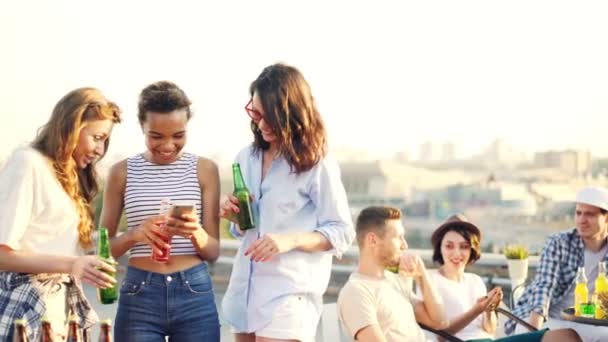 Radosny koleżanki są za pomocą smartfona i gawędząc gospodarstwa butelki podczas imprezy na dachu. Social media, młodych ludzi i koncepcji nowoczesnych technologii. — Wideo stockowe
