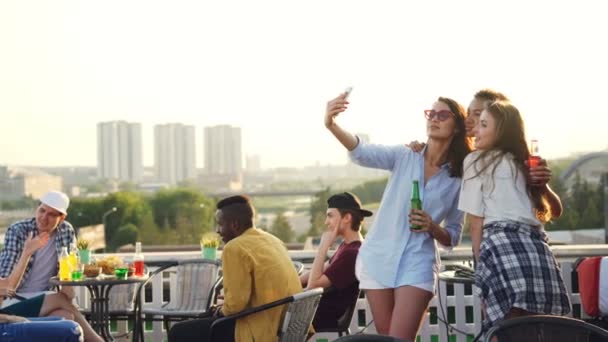 Mujeres jóvenes juguetonas se están tomando selfie con el teléfono inteligente haciendo caras divertidas que muestran la lengua y las orejas de conejo divertirse. Las niñas están usando ropa de moda y la celebración de botellas . — Vídeos de Stock