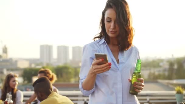 Pretty young woman is using smartphone, holding bottle and smiling during rooftop party with big city in background. Modern lifestyle and technology concept. — Stock Video