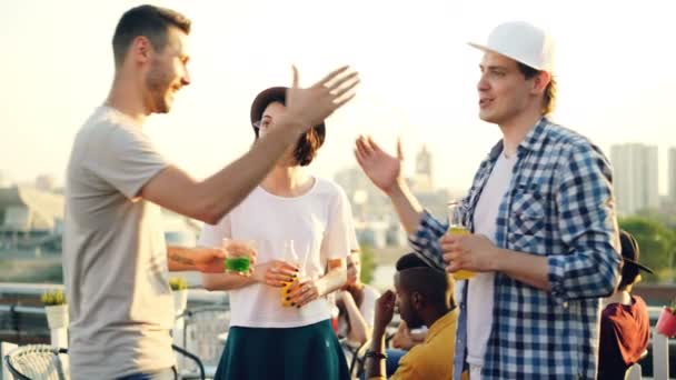 Attractive couple is talking then greeting young man at party on roof, men are shaking hands, woman is kissing her friend, they are clanging glasses and drinking. — Stock Video