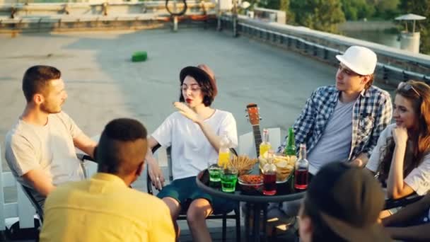 Amigos alegres están charlando y bebiendo limonada sentados en la azotea en un cálido día soleado con aperitivos y bebidas en la mesa. Concepto de verano, juventud y fiesta . — Vídeos de Stock