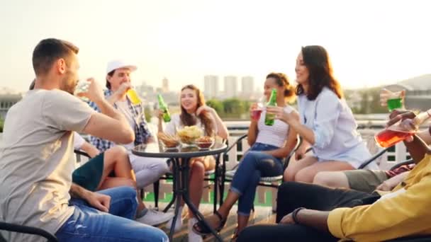 Los jóvenes alegres están celebrando las vacaciones en el techo bebiendo y tintineando botellas y vasos y luego hablando y riendo. Hermosa vista de la ciudad moderna está en el fondo . — Vídeos de Stock