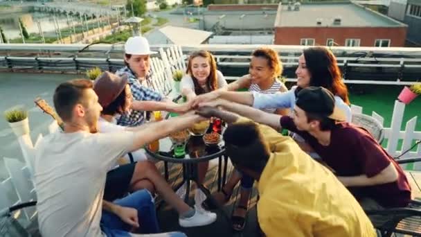 Amigos alegres estão colocando as mãos juntas, em seguida, levantando no ar sentado em círculo à mesa no telhado e beber. Conceito de integridade, festa e celebração . — Vídeo de Stock