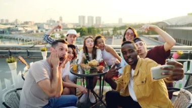 Genç Afrikalı-Amerikalı adam akıllı telefon tutarak ve onun arkadaşları çok etnik gruptan oluşan grup şişe ve çatı eğleniyor gözlük tutan selfie alarak.