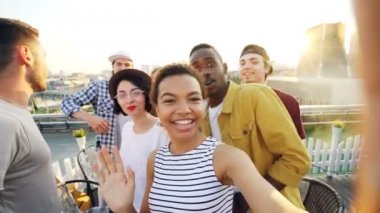 Neşeli Afro-Amerikan kız selfie ayar kamera ardından arkadaşları, poz ve çatı partide gülüyor görmek alıyor. Modern şehir arka planda görünür.