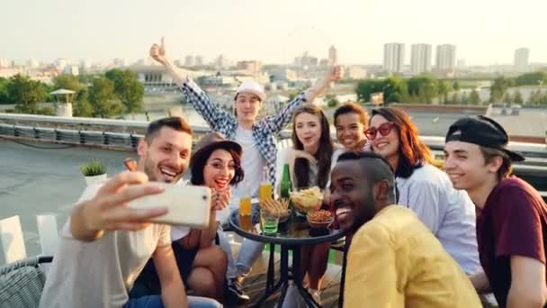 Giovani uomini e donne felici si stanno facendo selfie con lo smartphone in posa, ridendo e facendo gesti di mano celebrando la vacanza sul tetto a tavola con cibo e bevande . — Video Stock