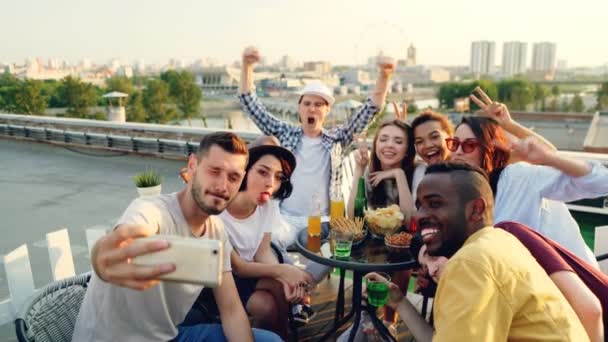 Cara barbudo está segurando smartphone e tirar selfie com amigos que estão posando com rostos engraçados e gestos de mão sentados à mesa no telhado se divertindo . — Vídeo de Stock