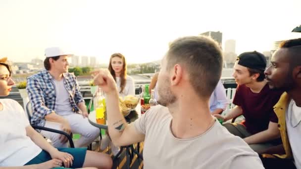 Point de vue prise de vue d'un jeune homme barbu regardant la caméra, touchant l'écran et ajustant la caméra, puis prenant selfie avec des amis assis sur le toit à table . — Video