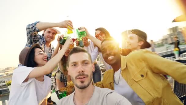Point of view shot of handsome guy holding camera and taking selfie while his friends are clinking glasses, having fun, touching his hair and ears and laughing. — Stock Video