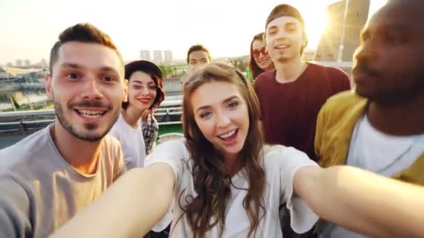 Jovem atraente está tomando selfie com amigos no telhado, menina está segurando câmera e posando enquanto seus companheiros estão se divertindo fazendo rostos engraçados e gestos . — Vídeo de Stock