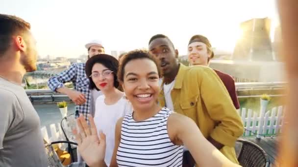 Menina afro-americana alegre está tomando selfie câmera de ajuste, em seguida, chamando seus amigos, posando e rindo de festa no telhado. Cidade moderna é visível no fundo . — Vídeo de Stock