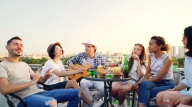 Süre arkadaşları şarkı, içme ve çatı masaya oturma gülüyorsun yakışıklı genç gitar çalıyor. Müzik, eğlence ve yaz aylarında konsept.
