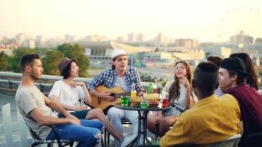 Çok ırklı bir grup genç insan, Genç adam gitar çalarken çatıda bir dairede oturan şarkı şarkıdır. Yiyecek ve içecekler ve güzel bir şehir olan tablo görülür.