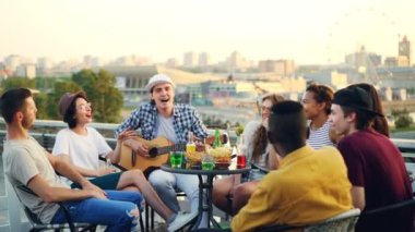 Neşeli müzisyen arkadaşlarıyla şarkı ve çatı masada oturan gülüyor gitar çalıyor. Eğlence, müzik aletleri ve eğlence kavramı.