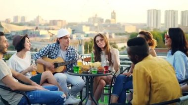 Mutlu arkadaşları şarkı, gülen ve çatı partisinde bira ve meşrubat içme iken erkek gitarist gitar çalıyor. Modern şehir arka planda görünür.