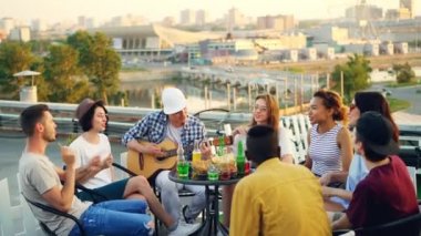 Gençler gitar çalmak çatıda şarkı, sohbet ve masada açık havada oturma gülüyor eğleniyor musun. Sevinç, müzik, gençlik ve dostluk kavramı.