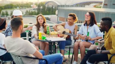 Afro-Amerikan Kız arkadaş çok ırklı grubu şarkı süre ve hareketli el çatıda eğleniyor gitar çalıyor. Tablo yiyecek ve içecek ile görünür.