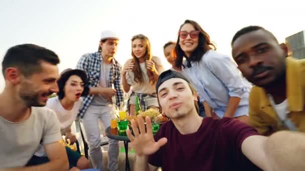 Punto de vista de la toma de joven alegre ajuste de la cámara y luego llamar a sus amigos para tomar selfie con bebidas en la fiesta en el techo, la gente se ríe y posando en el día soleado . — Vídeo de stock