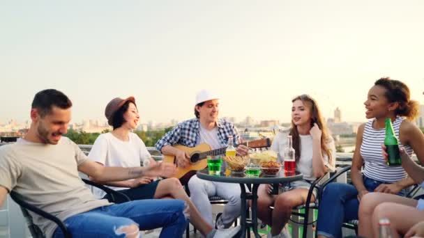 Chico creativo está tocando la guitarra y sus amigos multiétnicos están cantando botellas con cerveza y refrescos sentados en círculo en la mesa en el techo . — Vídeo de stock