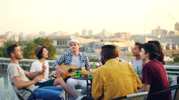 Students are having fun on rooftop playing the guitar, singing and dancing sitting at table on rooftop enjoying free time. Modern lifestyle and musical instruments concept. — Stock Video