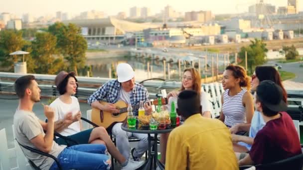 Os jovens estão se divertindo no telhado tocando guitarra, cantando, conversando e rindo sentados à mesa ao ar livre. Alegria, música, juventude e conceito de amizade . — Vídeo de Stock