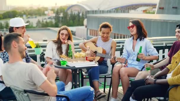 Atractiva chica afroamericana está tocando la guitarra, sus amigos están cantando, bebiendo y comiendo en la mesa en el techo disfrutando de fin de semana y cálido día soleado . — Vídeos de Stock