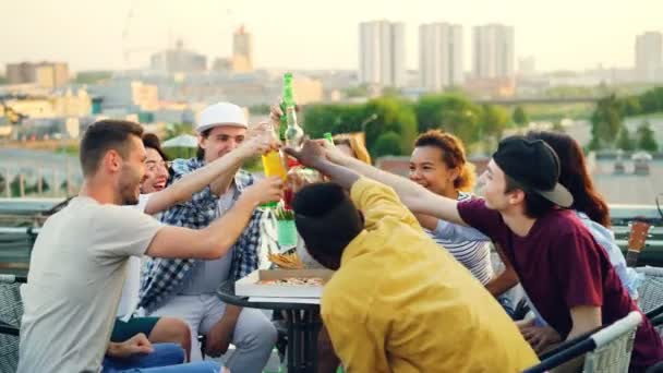 Os jovens são brindar, clinking garrafas e beber cerveja e refrigerantes, comer lanches, conversar e rir durante a festa no telhado no verão . — Vídeo de Stock