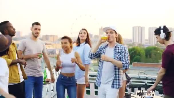 La juventud moderna está bailando en la azotea disfrutando de la música mientras su amigo DJ se mezcla trabajando con el equipo que lleva auriculares. Fiesta, celebración y concepto divertido . — Vídeo de stock