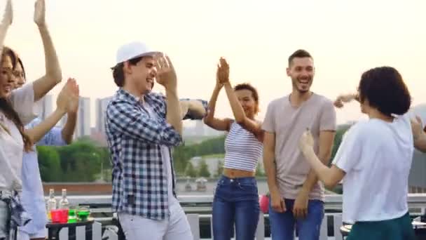Hombre joven guapo con gorra moderna está bailando con amigos en la fiesta en la azotea, hombres y mujeres se mueven y aplaudiendo las manos disfrutando del verano y el ocio . — Vídeos de Stock