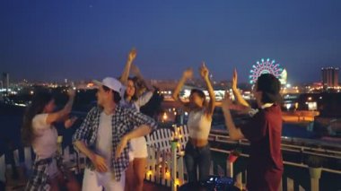 Neşeli Dj mikser ile gençler kızlar müzik zevk çatı partide çalışıyor ve çocuklar yükselterek eller dans ediyor. Güzel gece şehir arka planda görülmektedir.