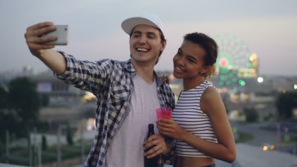 Alegre pareja multiétnica Caucásico joven y chica de raza mixta están tomando selfie en la azotea usando teléfono inteligente, riendo y sosteniendo bebidas . — Vídeos de Stock