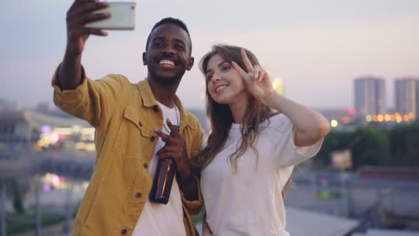 Casal multirracial atraente está tomando selfie com smartphone na festa no telhado, homem e mulher estão segurando bebidas e posando com rostos engraçados com a cidade no fundo . — Vídeo de Stock