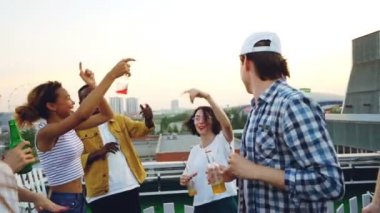 Gençlerin dans ve tatil kutluyor şişe tungur içecekler Çatıda rahatlatıcı ağır çekim. Güzel bir çatı görünümdür Metropolis arka planda.