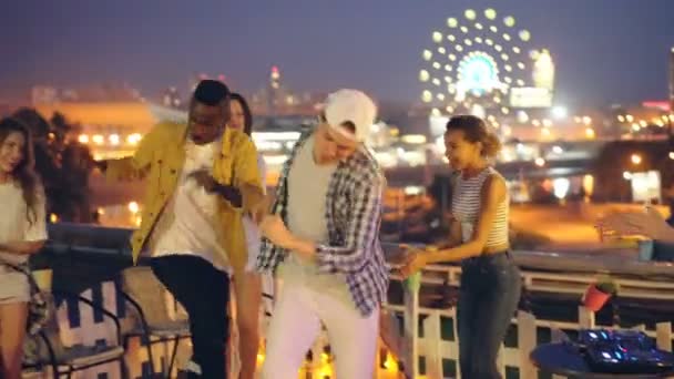 Joyful amigos meninas e caras estão dançando na festa à noite no telhado relaxante e rindo quando DJ está misturando música usando equipamentos especiais. Amizade e conceito divertido . — Vídeo de Stock